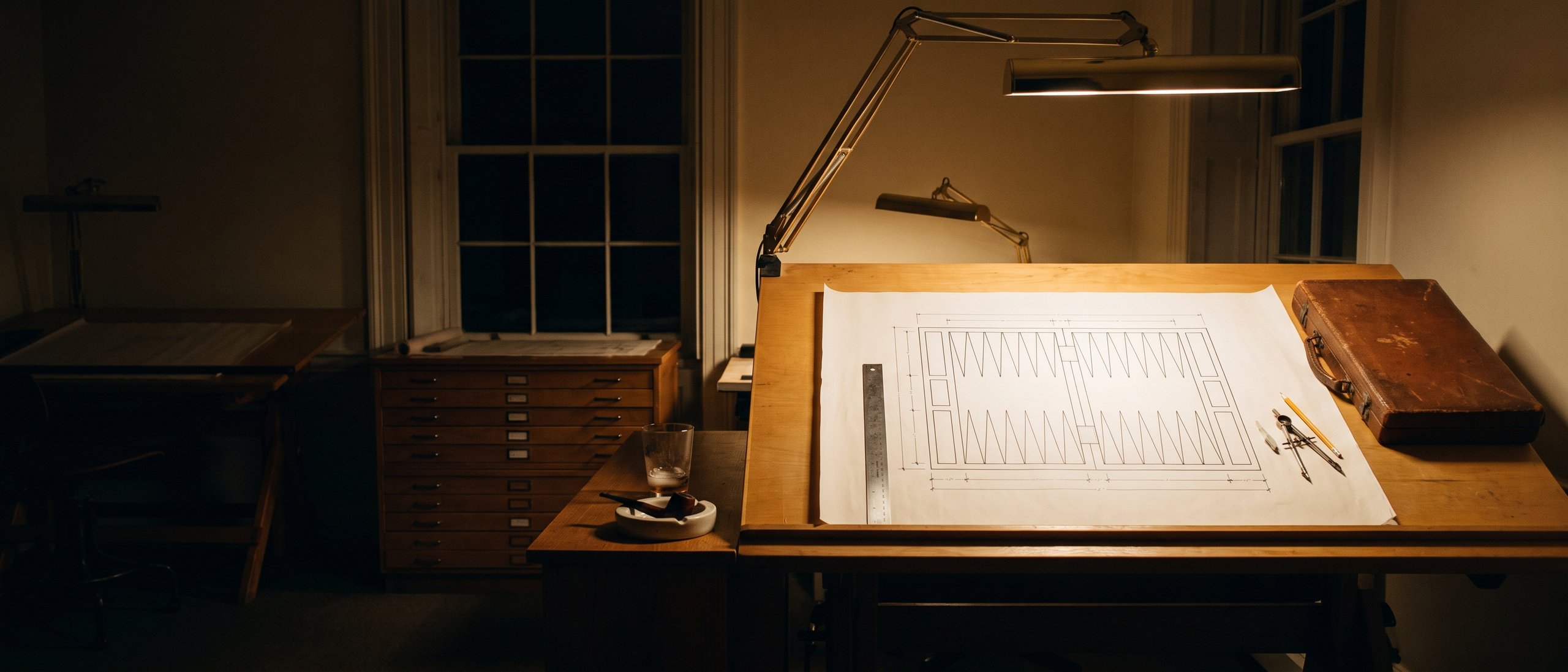 A technical drawing of a backgammon board on a drafting table under a lamp, with ruler and pencil